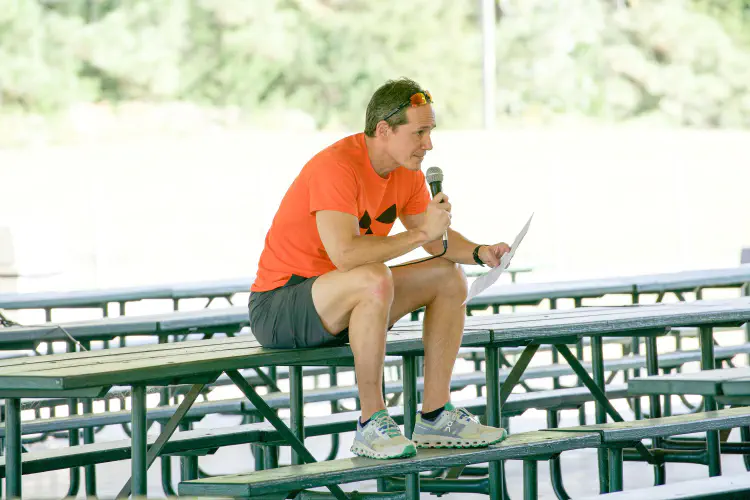 Man talking, holding microphone, sitting on bleachers